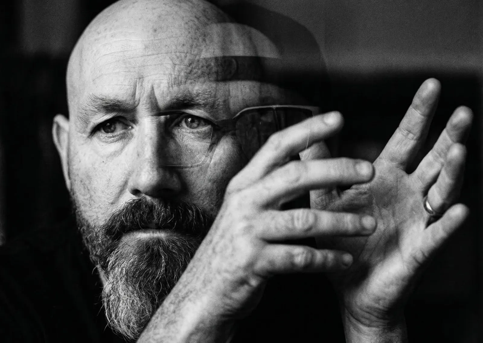 A moody, black and white double-exposure portrait of a bearded man with glasses. His face is sharp and contemplative, while a blurred overlay of his hands suggests rapid movement and physical intuition. The image has a gritty, film-grain texture, evoking a sense of psychological depth.