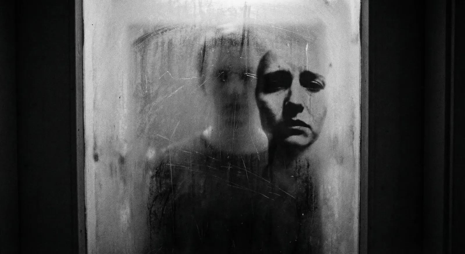 Moody black and white double exposure photography. A blurred, obscured human figure stands trapped behind heavily scratched frosted glass, while a sharp, dark silhouette of a different face is superimposed on the surface, symbolizing dissociation and the true self locked away.